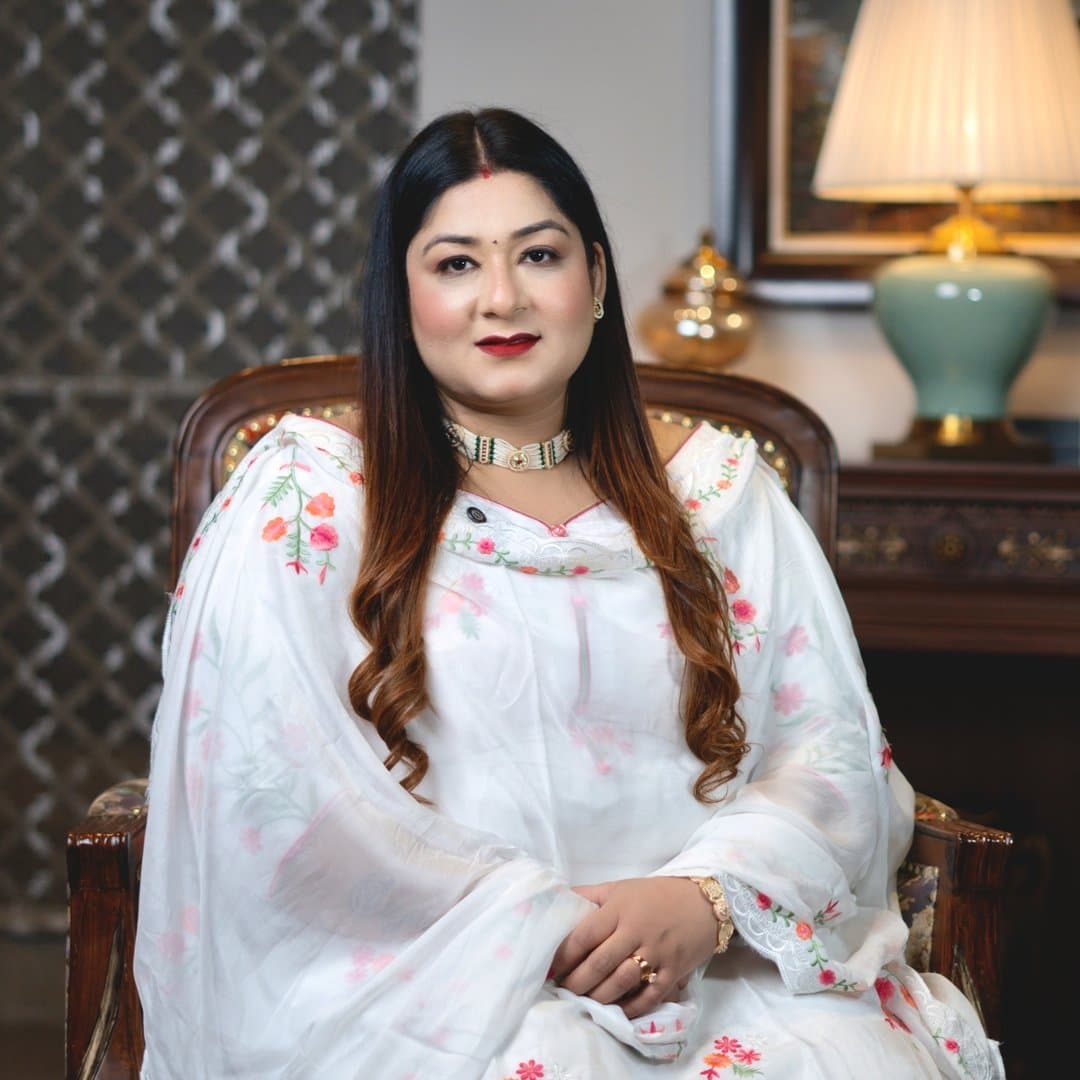 Komal Kalra seated in a carved wooden chair, wearing a white floral-embroidered suit, in a warmly lit room with a table lamp behind her.